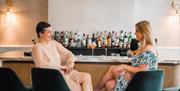 Two women sitting at a bar smiling at each other with cocktails behind them