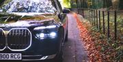 A black BMW car on a woodland road lined with orange fallen leaves and a black railing