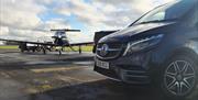 Side-view of a black Mercedes minivan parked at an airport. A jet airplane is parked in the background.