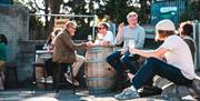 Guests at Saddle Goose's Cellar Door sat outside drinking from wine glasses on a sunny day. Some patrons are sat on bar stools while others are seated