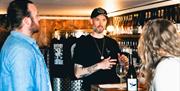 Adam from Saddle Goose, wearing a black t-shirt and cap, talks to two patrons at the bar with a wine glass and bottle in front of him. Shelves filled
