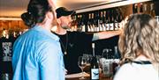 Adam at Saddle Goose, wearing a black t-shirt and cap, points at a row of wine bottles on a shelf while serving two customers.