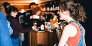 A person wearing dungarees drinks from a wine glass. Out of focus in the background, two members of bar staff can be seen serving a line of customers.