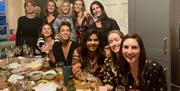A group of ten girls smiling and toasting along a long wooden table with empty bowls and wine glasses on the table.