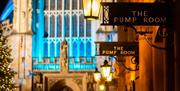 Signs for The Pump House at night, lit by lanterns. Bath Abbey and a decorated tree are both visible out-of-focus in the background.
