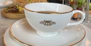 A white china with gold rim Hope & Glory branded tea cup and saucer with food in background at The Abbey Tea Bar, Bath