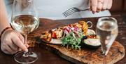 A woman sitting at a table eating a kebab and pitta wrap with dips alongside glasses of wine