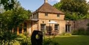 A sandstone country house with tiled pitched roof, a table, chairs and white parasol outside next to a garden with a sculpture in (Barbara Hepworth in