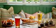 A pint of cider, a pint of ale, and fish and chips on a wooden pub table with a bench with cushions on it in the background.