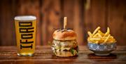 A trio of a pint of cider, a burger, and a bowl of chips on a wooden bench.
