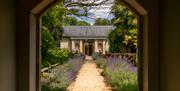 A stone archway leading down a lavender lined path to a sandstone outhouse with a slate pitched roof and wooden door. The entrance to the spa.