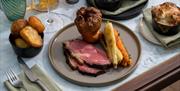 A plate of pink roast beef with parsnips, carrots and a large Yorkshire pudding on it. There are small round side bowls of roast potatoes, cauliflower