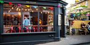 The exterior of The Village cafe and bar, with several people seated in a terrace to the right hand side. Multi-coloured bunting lines the top of the