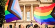 A photograph of the front of the Holburne Museum with a crowd of people in front. Two large progress rainbow flags fly in the foreground.
