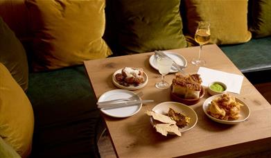 Several plates of food on a square table, next to a bench with green and yellow cushions