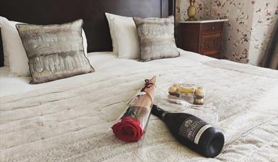 A bottle of Prosecco, a bouquet of flowers, and a box of chocolates on top of a bed.