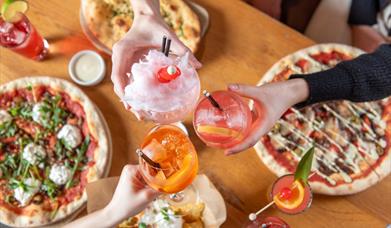 A table of pizzas and various side dishes beneath three hands clinking drinks together