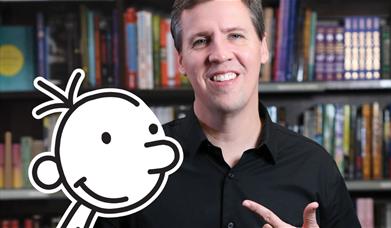 Author Jeff Kinney stands in front of a bookshelf, with a cut-out of the character Greg Heffley in front of him.