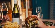 A hand holding a burger with a bowl of chips and a wine bottle in the background at Green Park Brasserie.