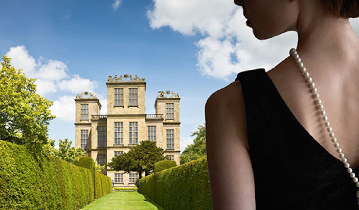 Woman in black dress stood in front of a garden filled with tall green hedges and a large manor house