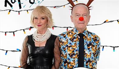 A photo of Toyah and Robert in front of a white background. Behind them are strings of Christmas lights. Toyah is wearing a black leather dress with a