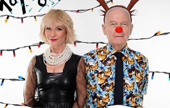 A photo of Toyah and Robert in front of a white background. Behind them are strings of Christmas lights. Toyah is wearing a black leather dress with a