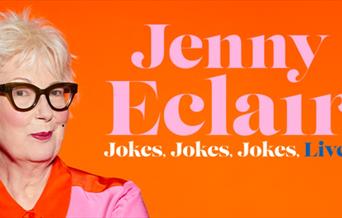 Jenny Eclair in a pink shirt, standing in front of a orange background.
