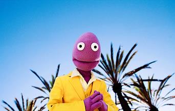 Randy Feltface in a yellow suit standing against a sunny background.