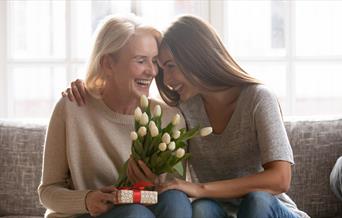 A mum and daughter sat on the sofa exchaning gifts and flowers
