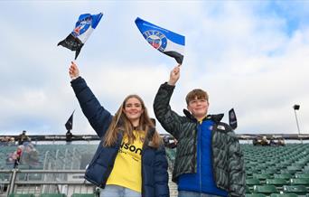 Two young Bath Rugby fans waving flags in the stands, showing support for their club during a match.