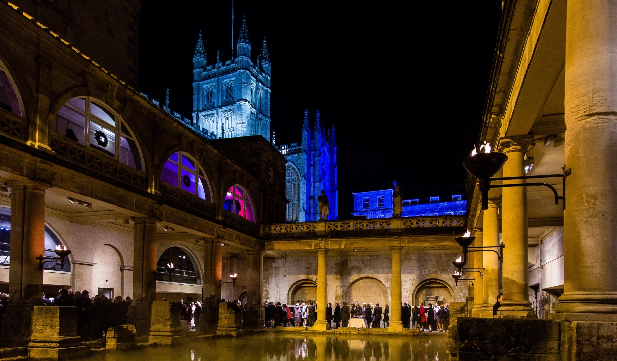 An image of The Great Bath at The Roman Baths at night with all the torches lit. The Bath Christmas lights can be seen illuminating Bath Abbey in the