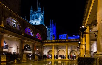 An image of The Great Bath at The Roman Baths at night with all the torches lit. The Bath Christmas lights can be seen illuminating Bath Abbey in the
