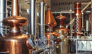 An image from Glendalough distillery from the still house. An assortment of shiny copper stills and stainless steel pipes. In the backgroud is the gle