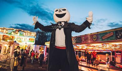 Night time shot of a spooky character in a black suit wearing a white pumpkin typr jack-o-lantern head. There is a fairground in the background.
