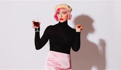 Photo of Tatty Macleod posing with a glass of red wine her hand. She has bright red lipstick and is wearing a light yellow beret, a black turtle neck 