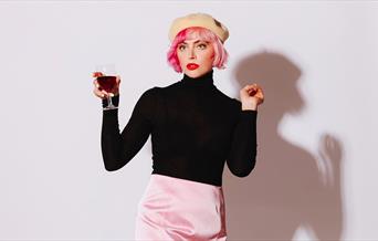 Photo of Tatty Macleod posing with a glass of red wine her hand. She has bright red lipstick and is wearing a light yellow beret, a black turtle neck