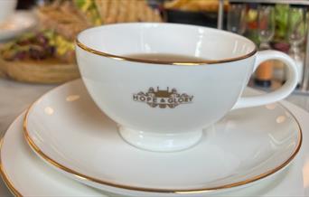 A white china with gold rim Hope & Glory branded tea cup and saucer with food in background at The Abbey Tea Bar, Bath