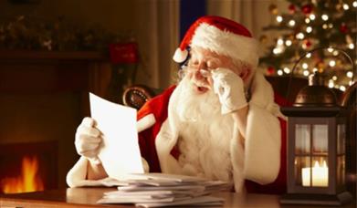 A man dressed as Father Christmas reading a Christmas present list at a table with a Christmas tree in the background 