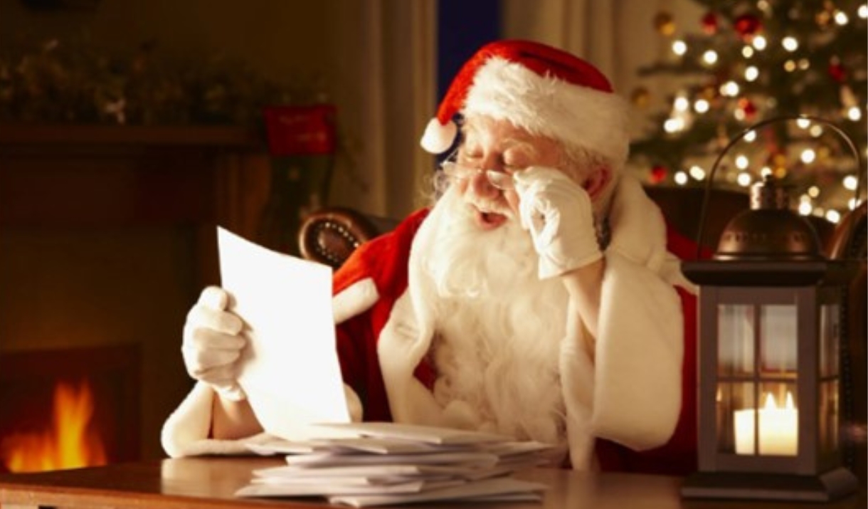 A man dressed as Father Christmas reading a Christmas present list at a table with a Christmas tree in the background