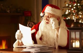 A man dressed as Father Christmas reading a Christmas present list at a table with a Christmas tree in the background