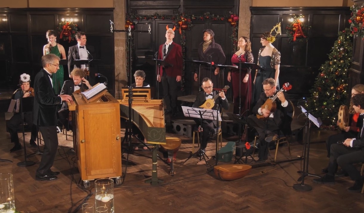 Small orchestra sat round a harpsichord with four singers standing behind them. All stood in a festive panelled room.