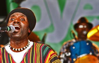 Two African musicians wearing colourful clothes - one signing in focus at the front and a drummer out of focus behind.