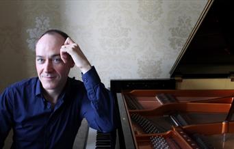 Leon McCawley sat sideways at a piano with his elbow up on the piano.